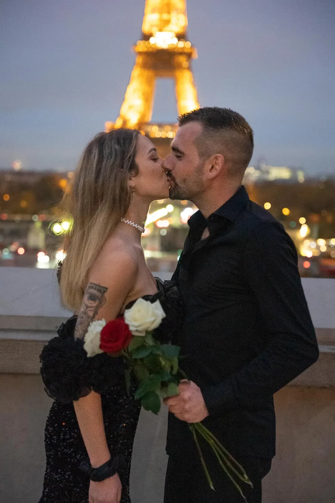 A couple kissing at dusk in front of the illuminated Eiffel Tower, holding a bouquet of red and white roses.