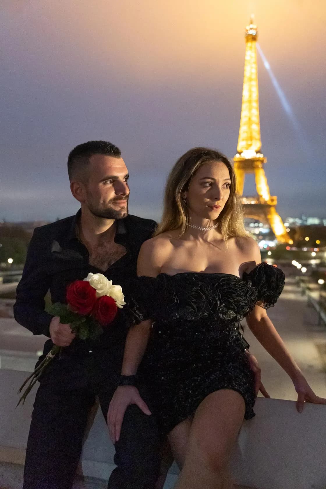 A couple dressed in black at dusk, roses in hand, in front of the illuminated golden Eiffel Tower