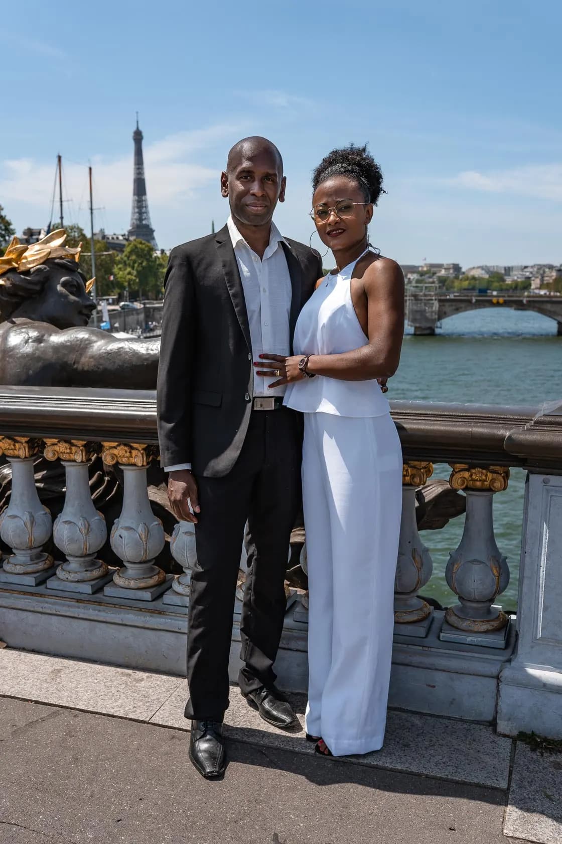 Elegant couple, one in a black suit and the other in a white jumpsuit, posing on the Pont Alexandre III, with the Eiffel Tower in the distance