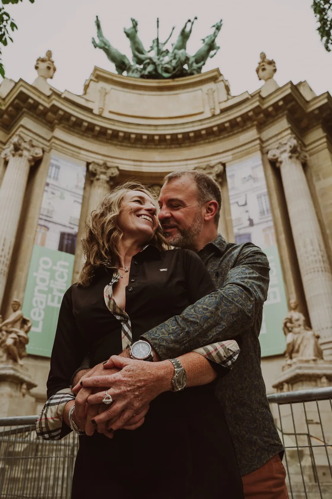 A couple embraces in front of the entrance to the Grand Palais in Paris, with the quadriga statue in the background.