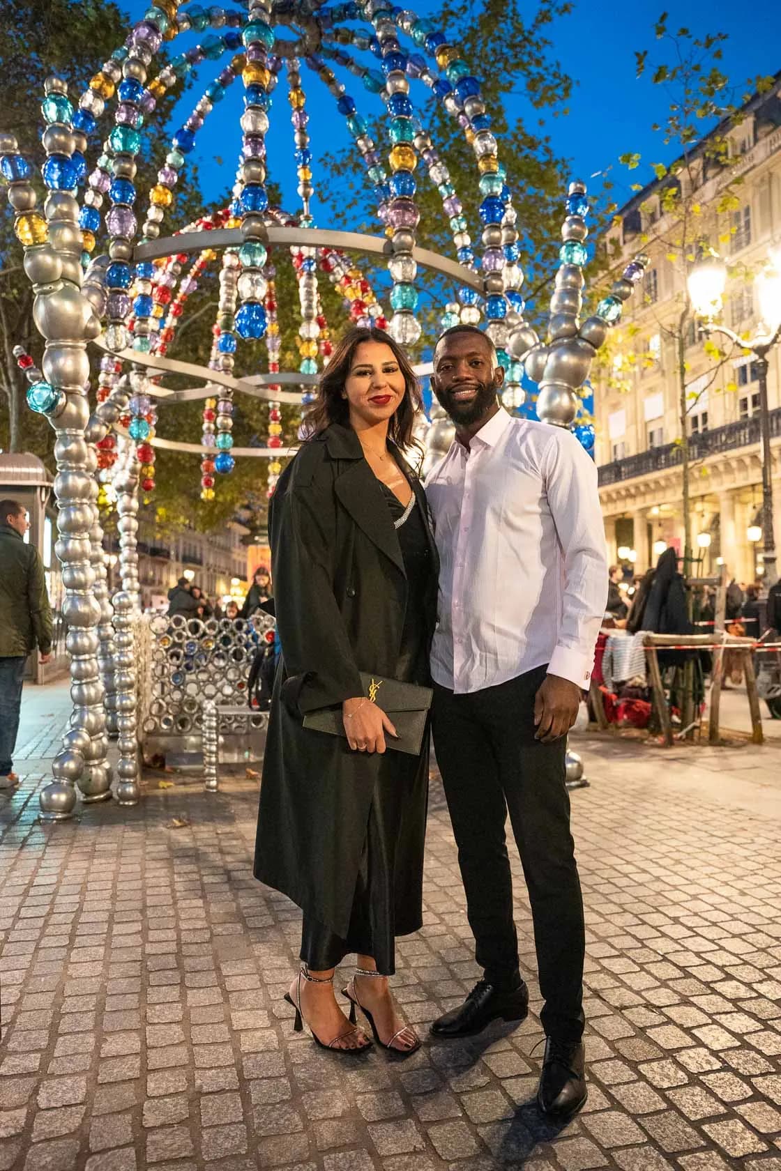 An elegant couple posing under a luminous arch of colorful beads, on a lively Parisian street in the evening