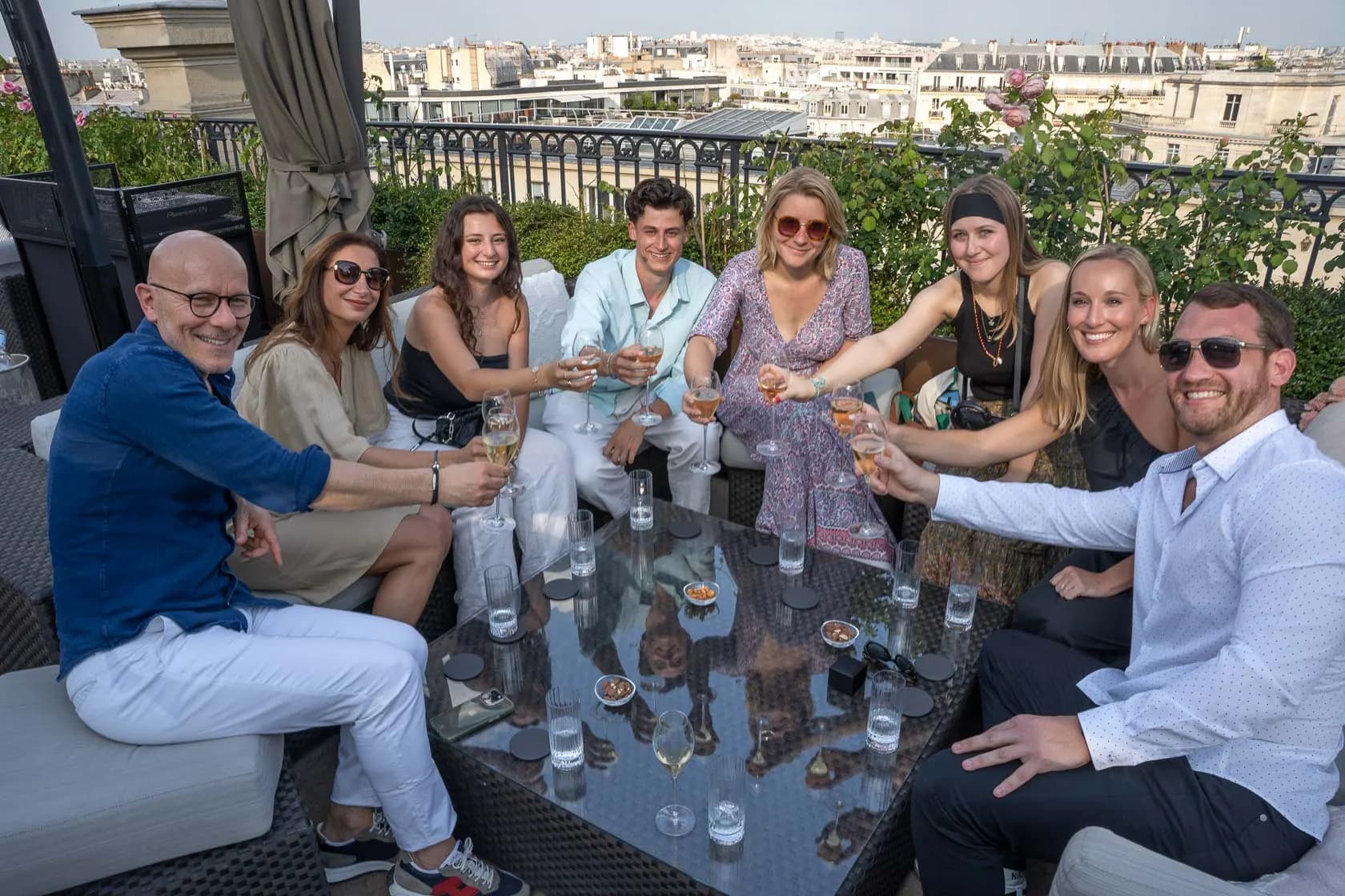 A group of friends and relatives toasting with champagne around a table on a terrace overlooking the rooftops of Paris at sunset.