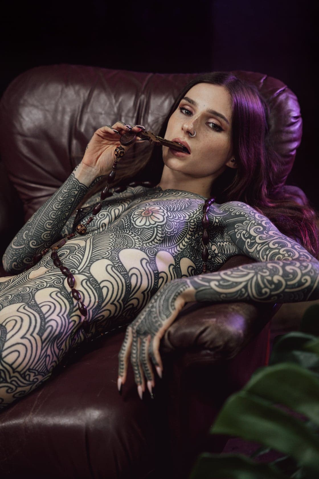 Portrait boudoir de Noémie Doragon nue, croquant un chapelet, allongée sur un canapé dans un décor coloré