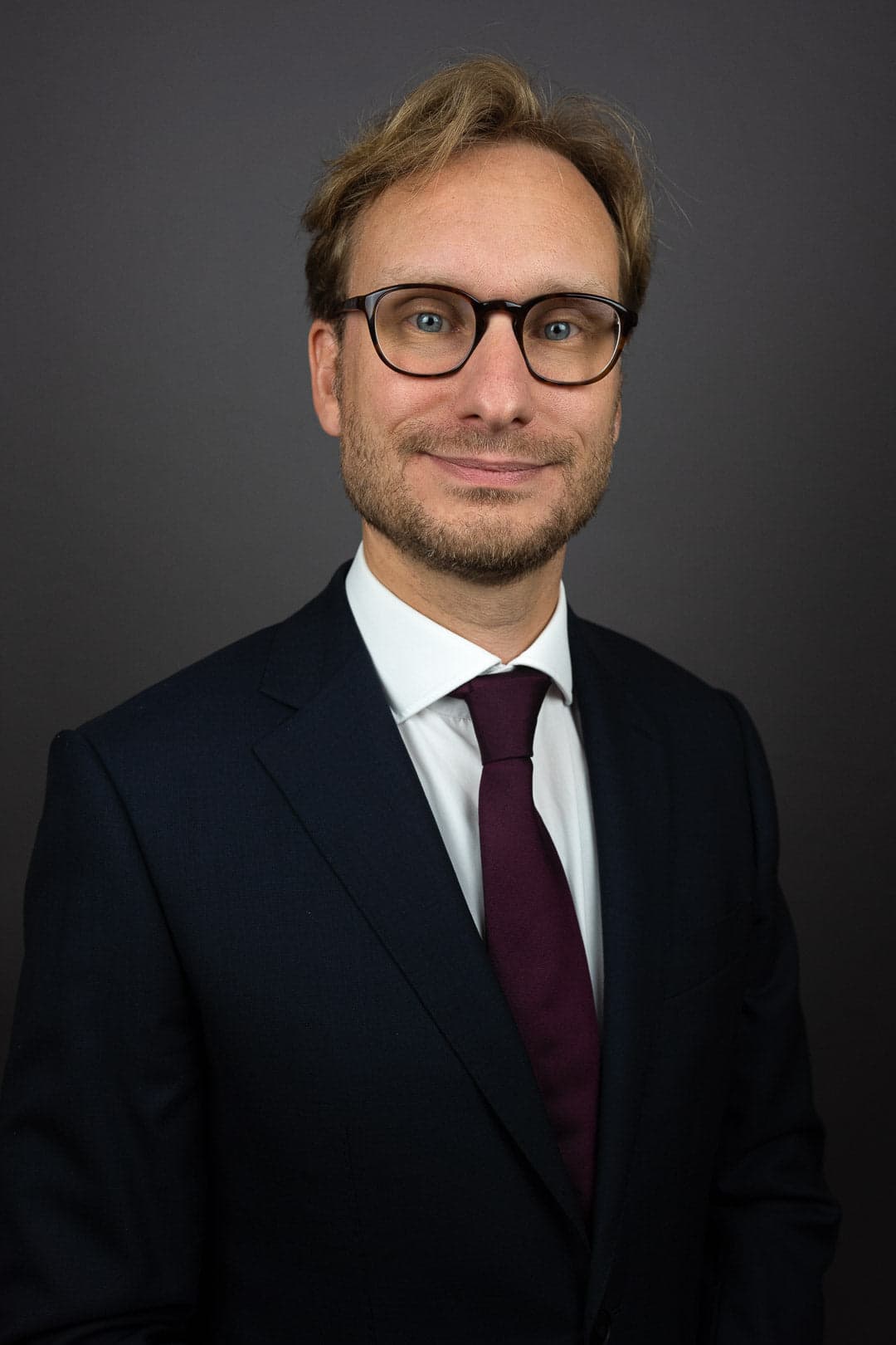 Portrait d'un homme aux cheveux blonds et aux yeux bleus portant des lunettes noires, un costume bleu marine, une chemise blanche et une cravate bordeaux