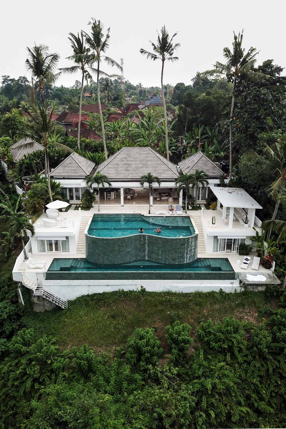 Vue aérienne d'une luxueuse villa blanche à Ubud, avec une grande piscine à débordement au design unique, entourée de végétation tropicale luxuriante et de palmiers