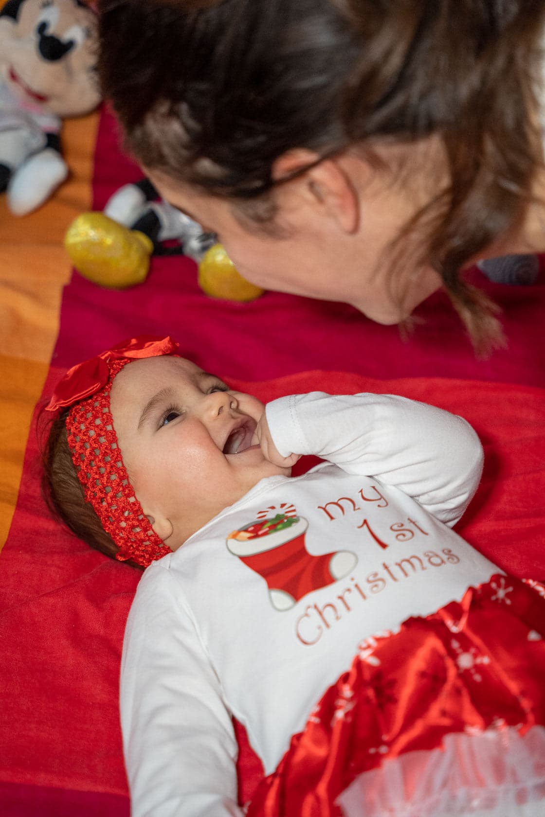 Bébé souriant en tenue de Noël 'My 1st Christmas' sur un lit, sa maman se penchant au-dessus