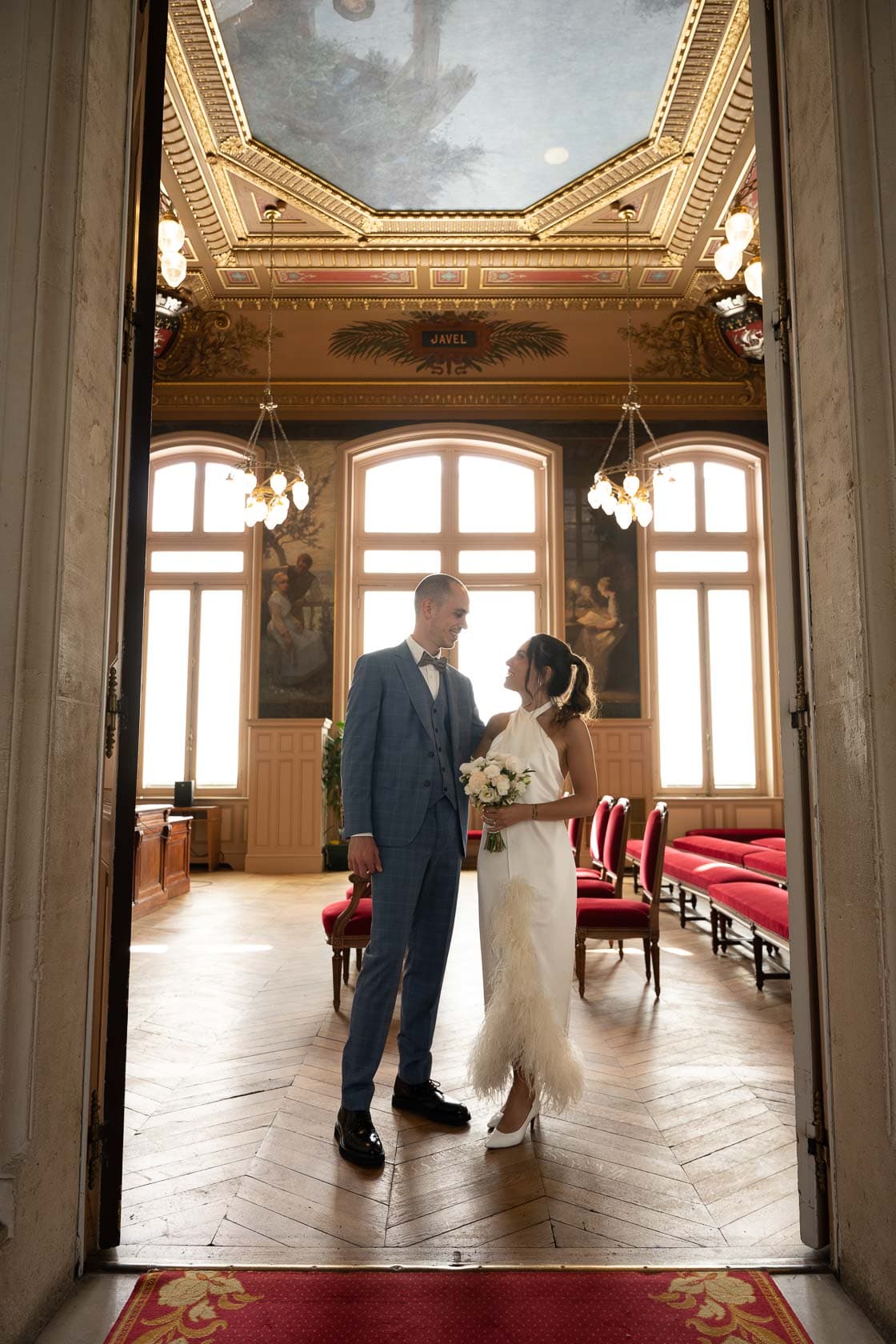 Les mariés échangeant un regard complice dans une salle majestueuse, illuminée par de grandes fenêtres