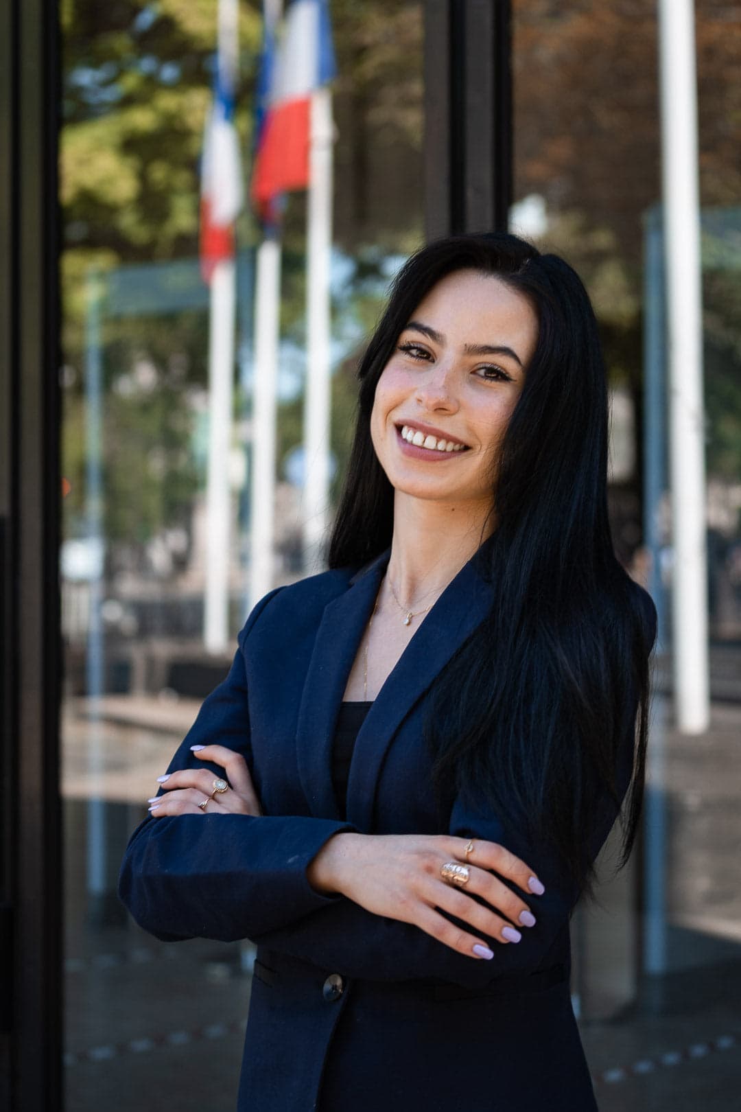Portrait d'une jeune femme souriante aux longs cheveux noirs, portant un blazer bleu marine, bras croisés devant un bâtiment avec des drapeaux français