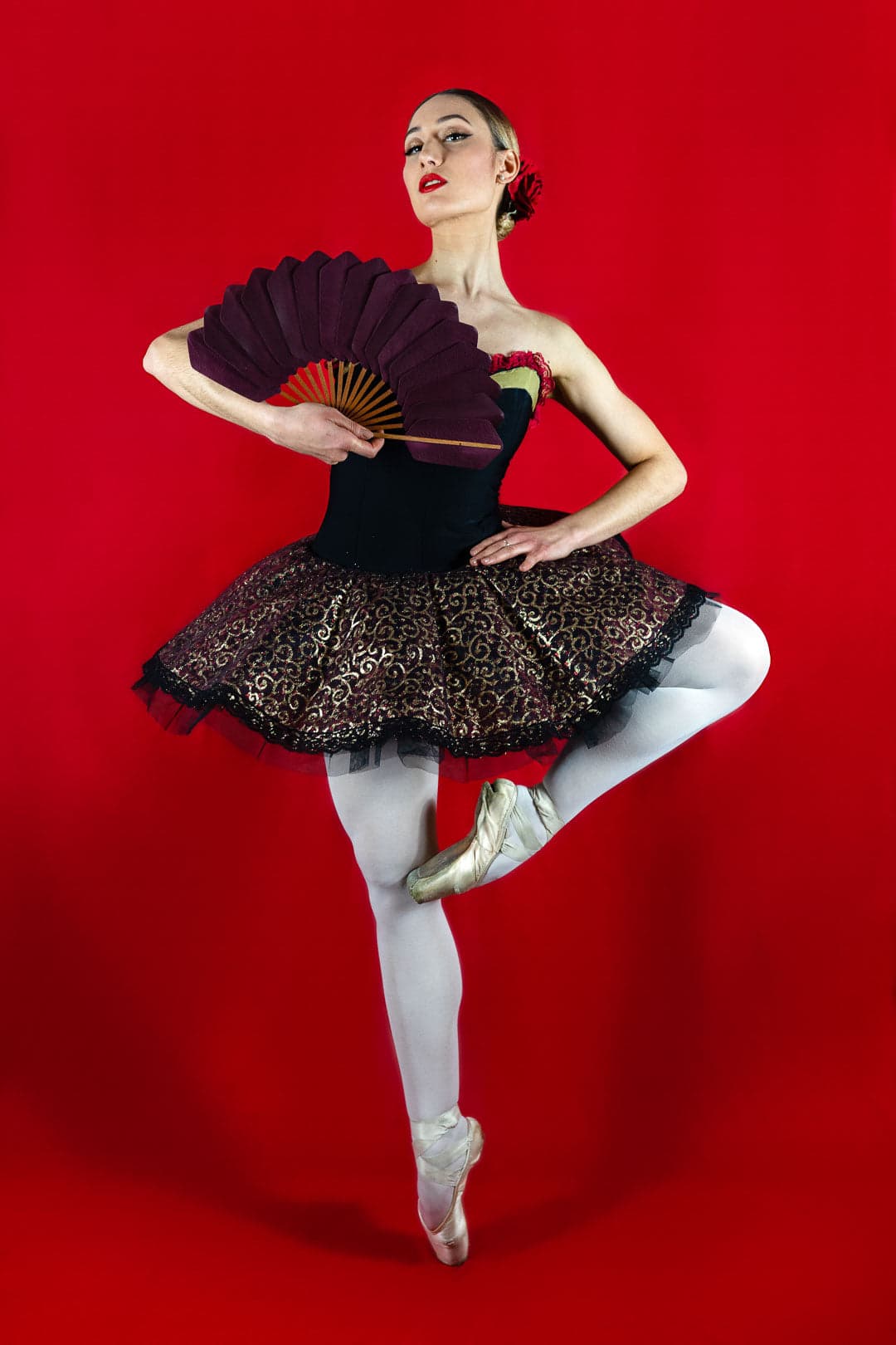 Ballerine en tutu noir et or, collants blancs et chaussons de danse, tenant un éventail bordeaux sur fond rouge