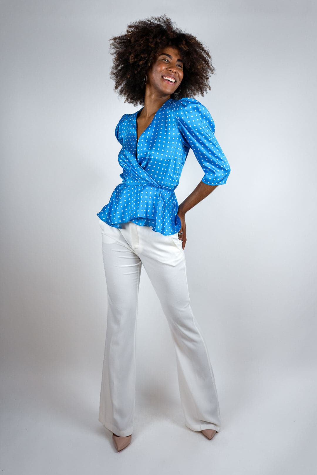 Femme souriante, peau foncée, cheveux afro volumineux, chemisier cache-cœur bleu à pois blancs, pantalon blanc évasé, studio sur fond gris clair