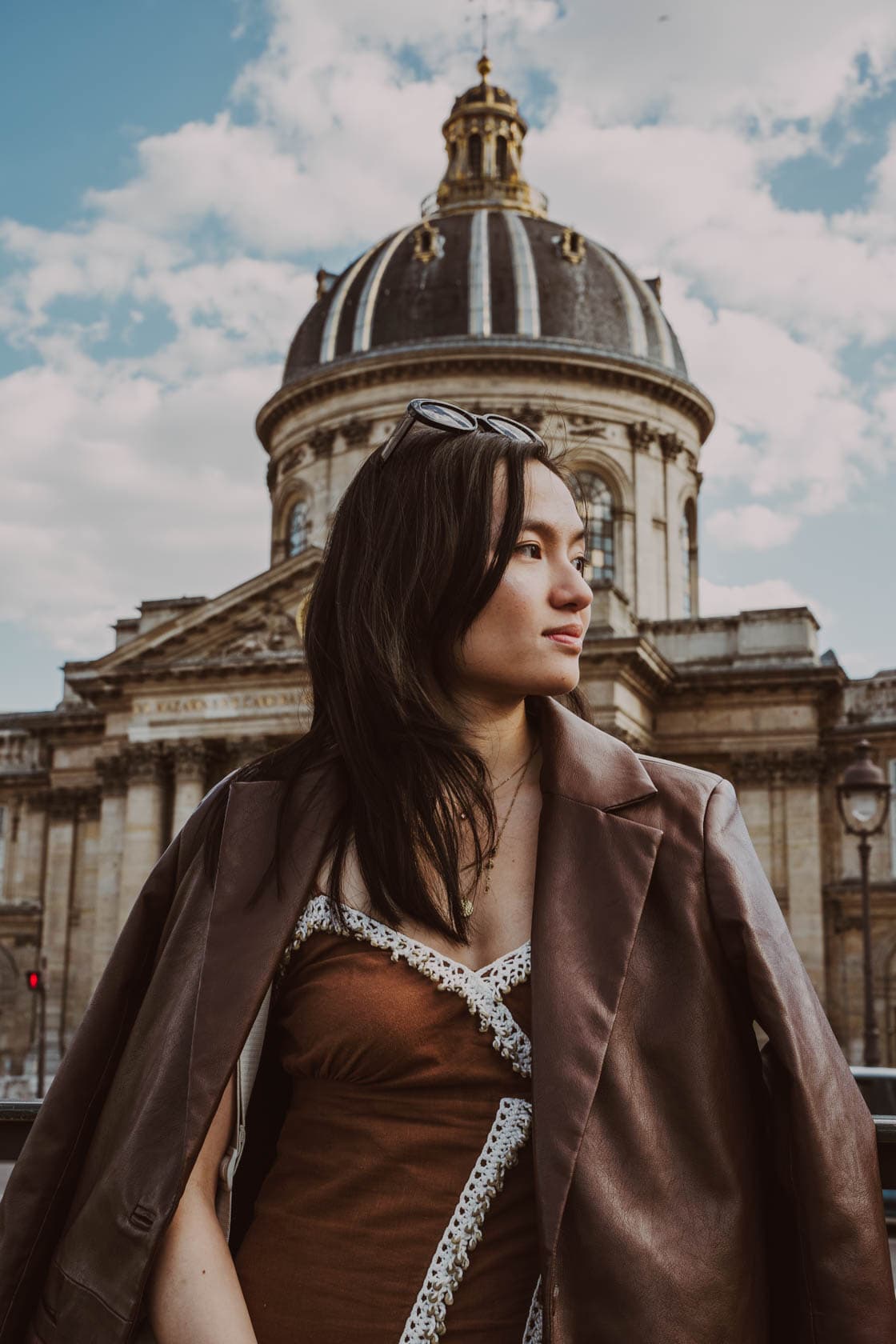 Jeune femme aux cheveux longs et ondulés, portant un blazer marron sur un haut clair avec de la dentelle, posant devant un grand dôme architectural