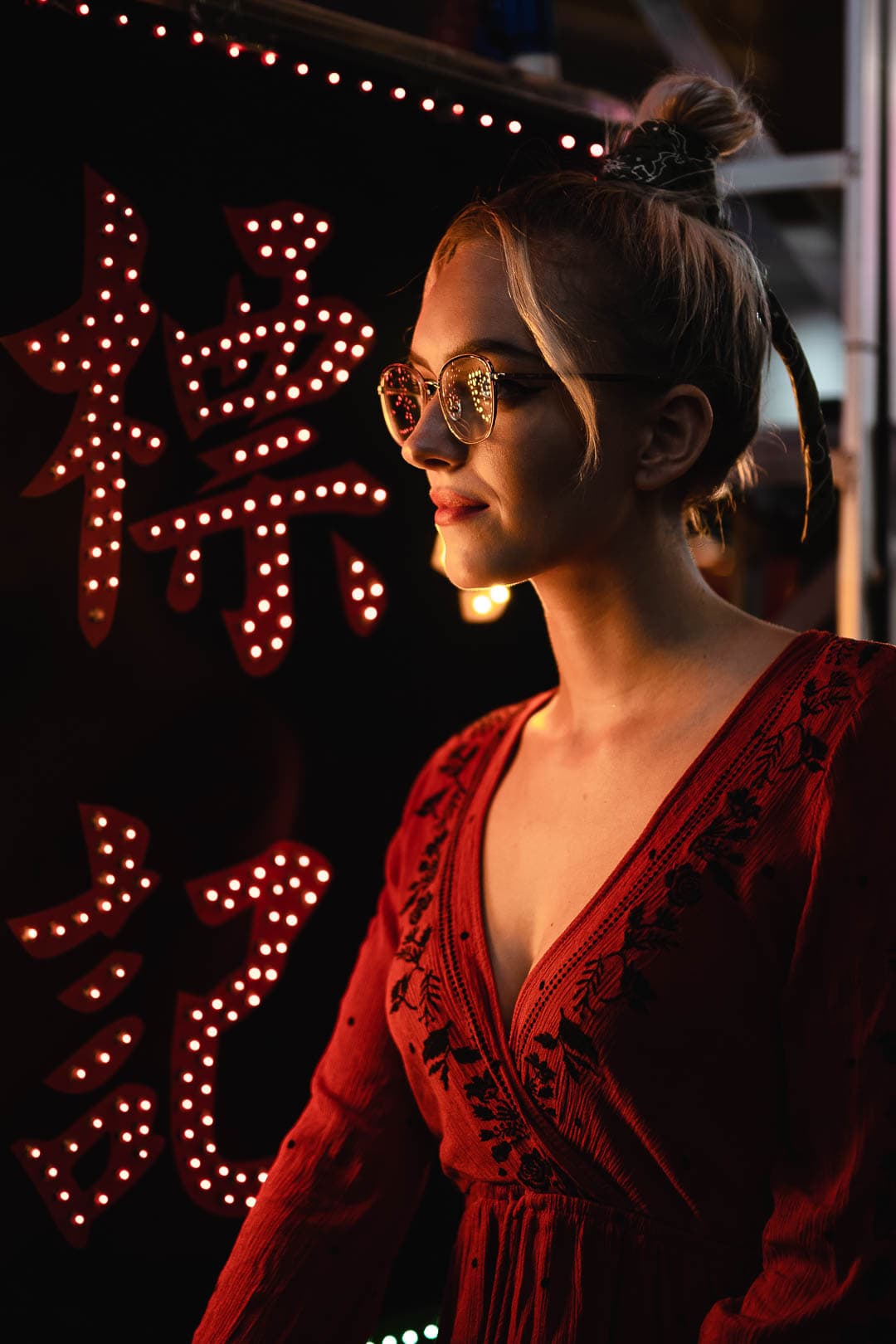 Portrait d'une jeune femme blonde portant des lunettes rondes, une robe bordeaux et posant près d'une enseigne lumineuse en caractères chinois