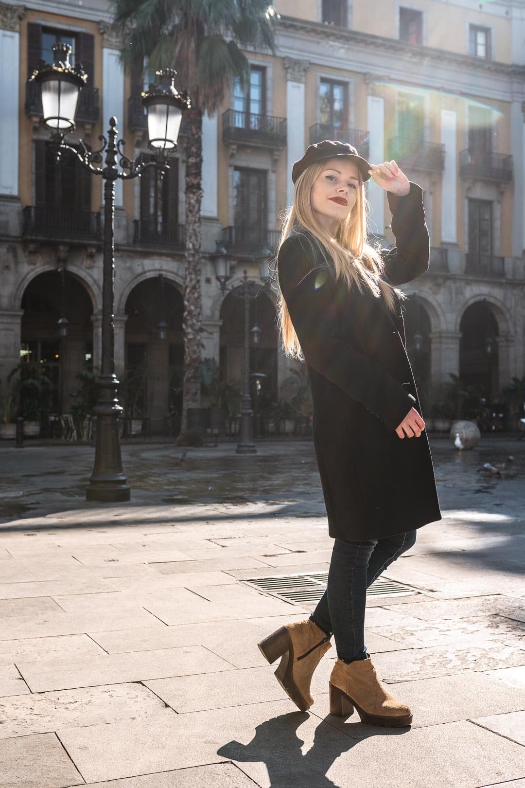 Femme blonde portant un manteau noir, des collants et des bottines à talons, posant sur la Plaça Reial à Barcelone, place à arcades avec ses lampadaires historiques