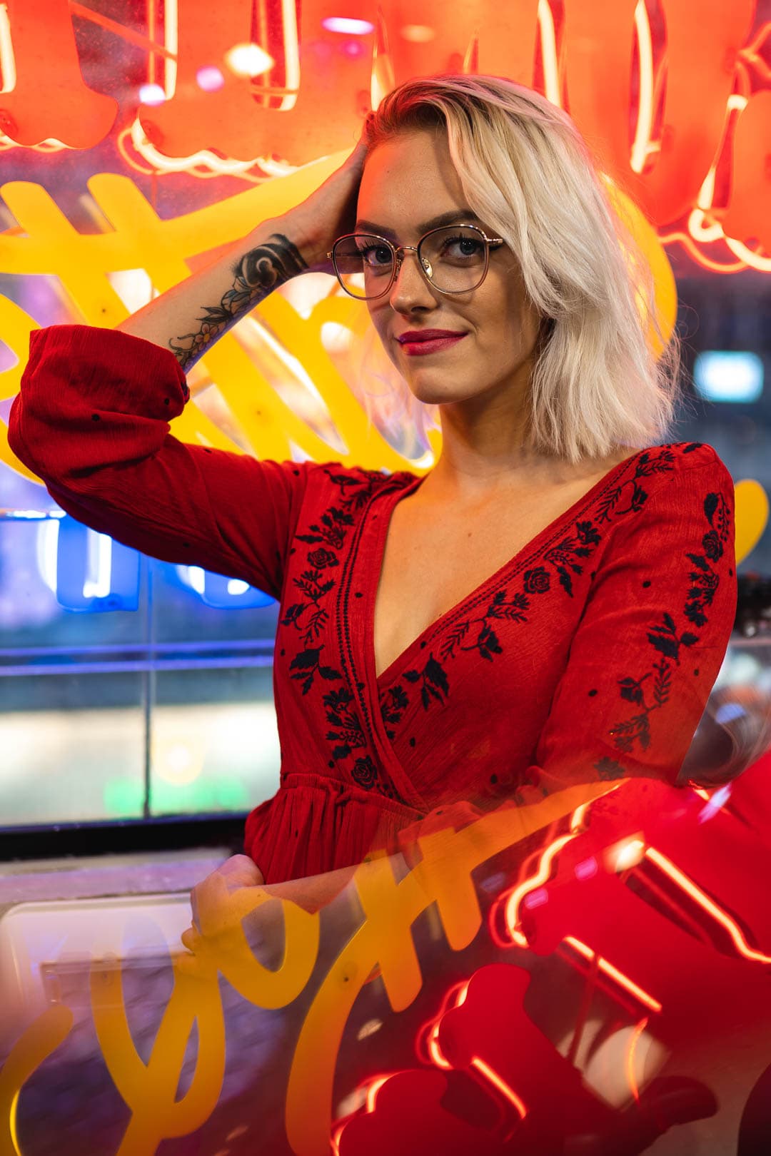 Femme blonde portant des lunettes et une robe rouge à motifs, posant devant une enseigne lumineuse colorée