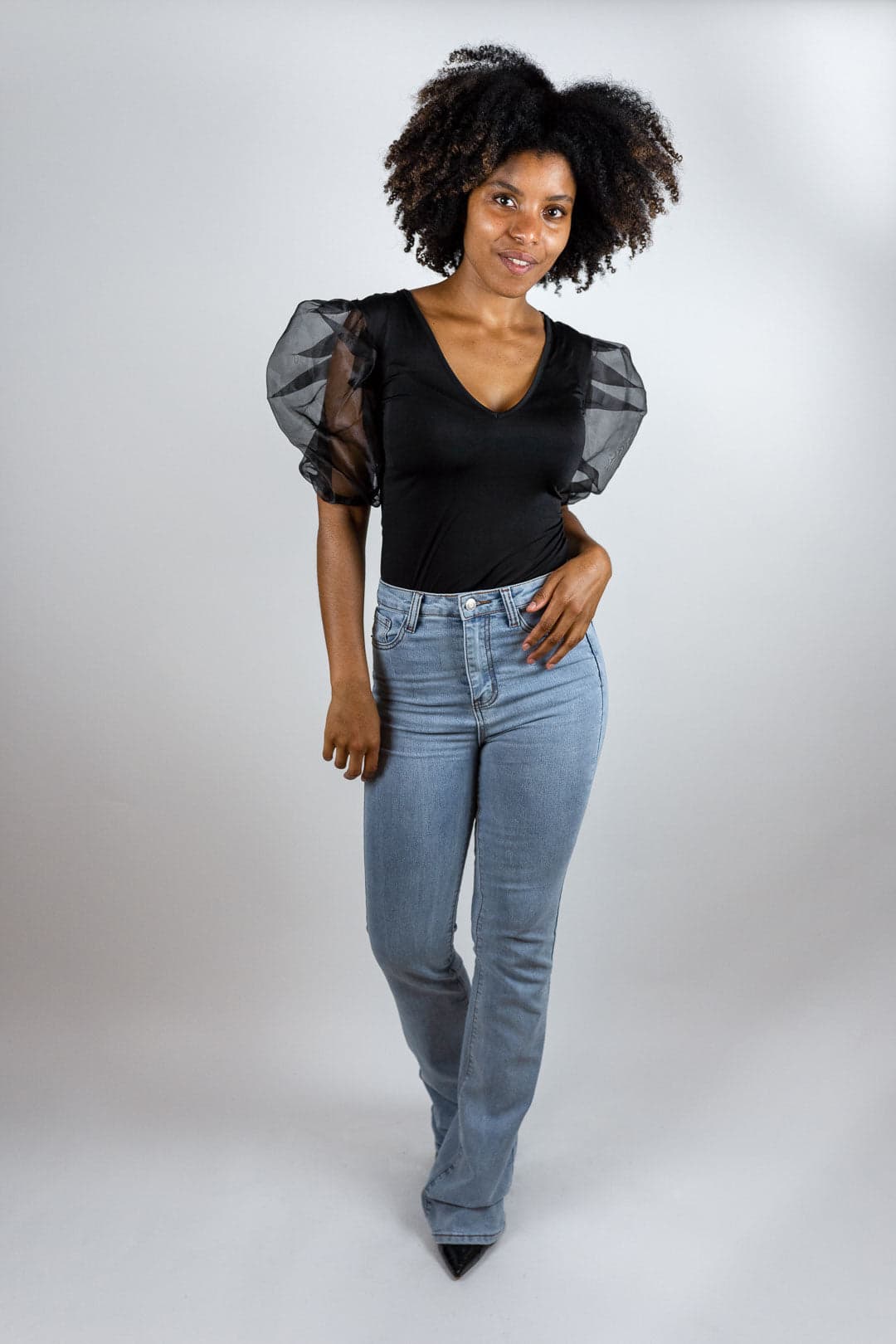 Femme souriante en body noir à manches bouffantes transparentes et jean bleu clair, studio sur fond gris clair.