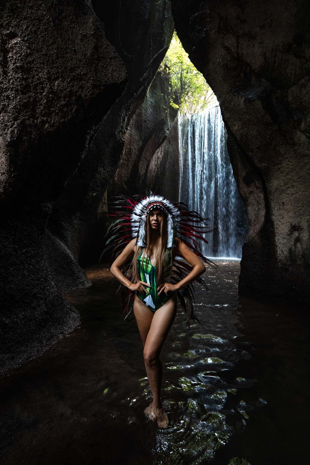 Femme portant une coiffe amérindienne et un maillot de bain, posant dans une grotte sombre avec une cascade en arrière-plan