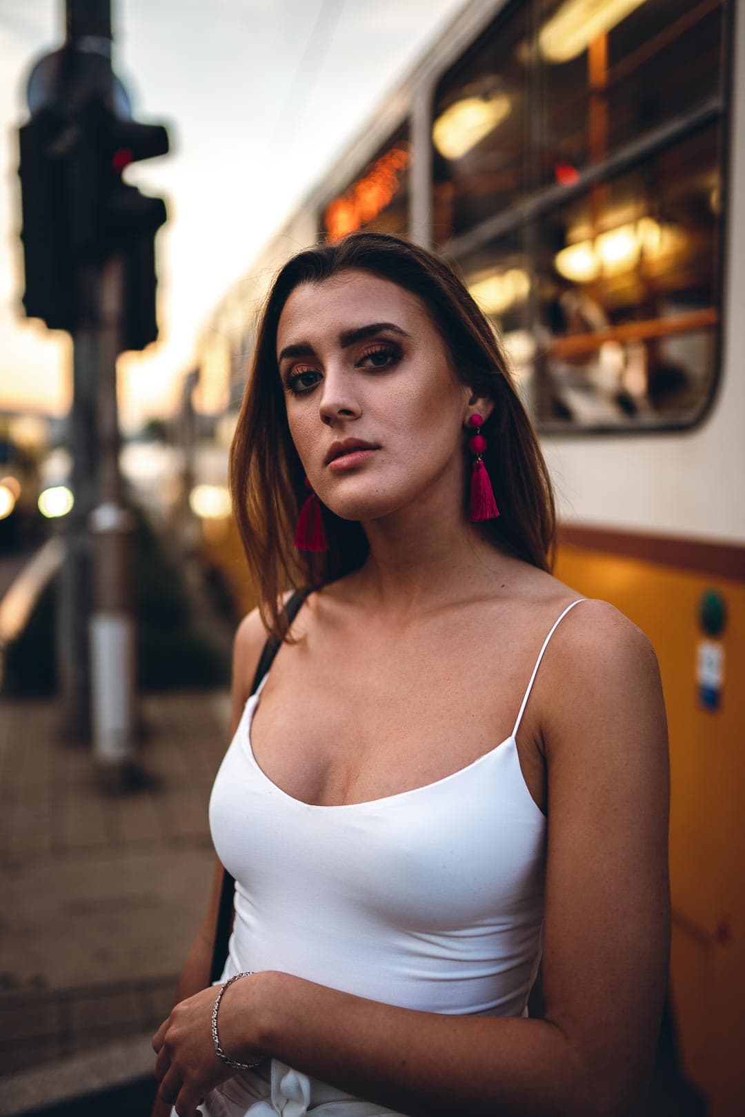 Portrait d'une jeune femme aux longs cheveux bruns, portant un débardeur blanc et de longues boucles d'oreilles rouges, regardant la caméra avec une expression sérieuse, un tramway flou de Budapest en arrière-plan
