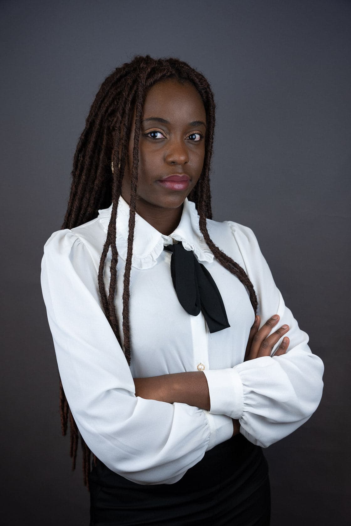 Portrait d'une jeune femme noire avec des dreadlocks, portant un chemisier blanc à col à volants et un nœud papillon noir, bras croisés