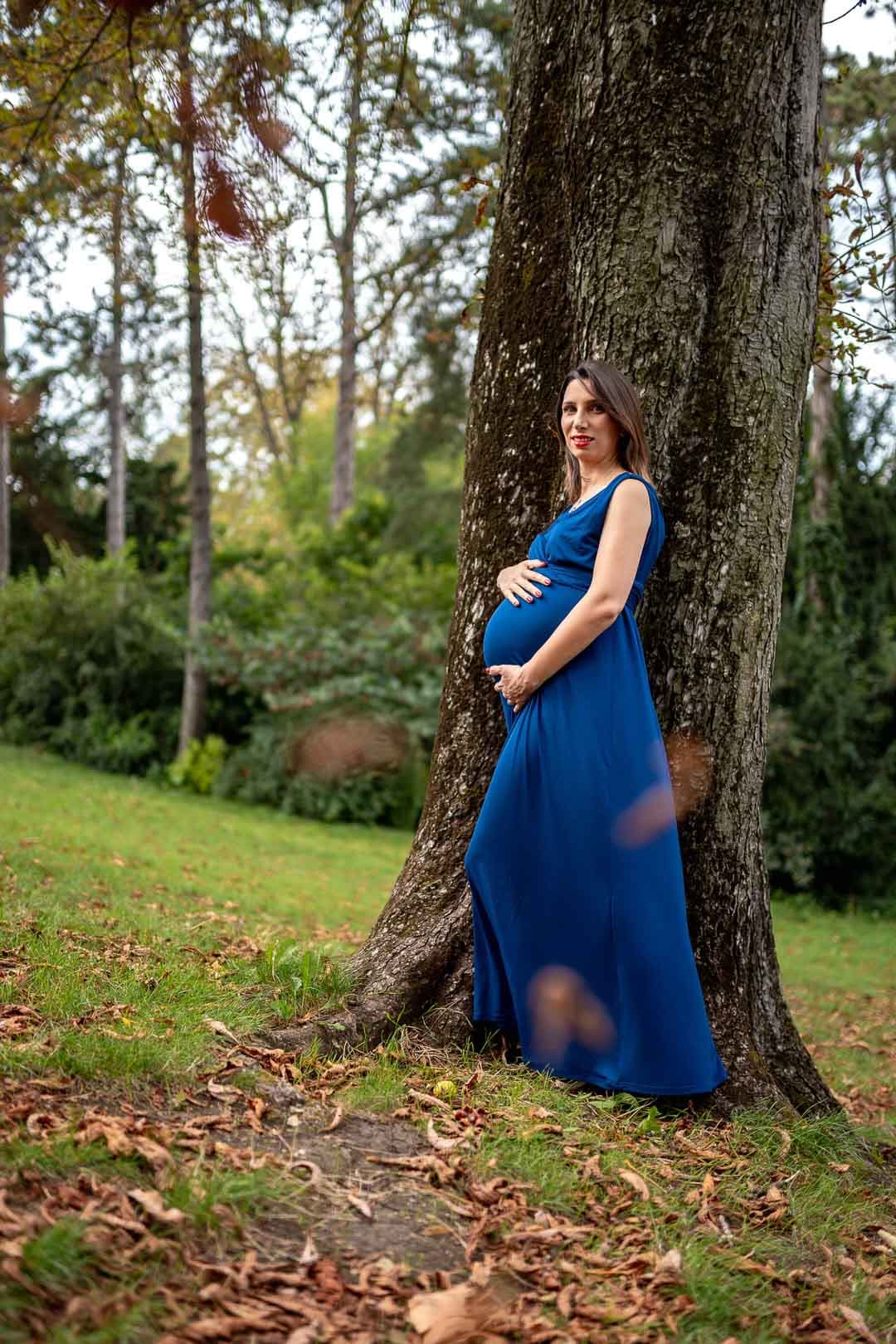 Femme enceinte en longue robe bleue posant près d'un grand arbre dans au parc Montsouris en automne