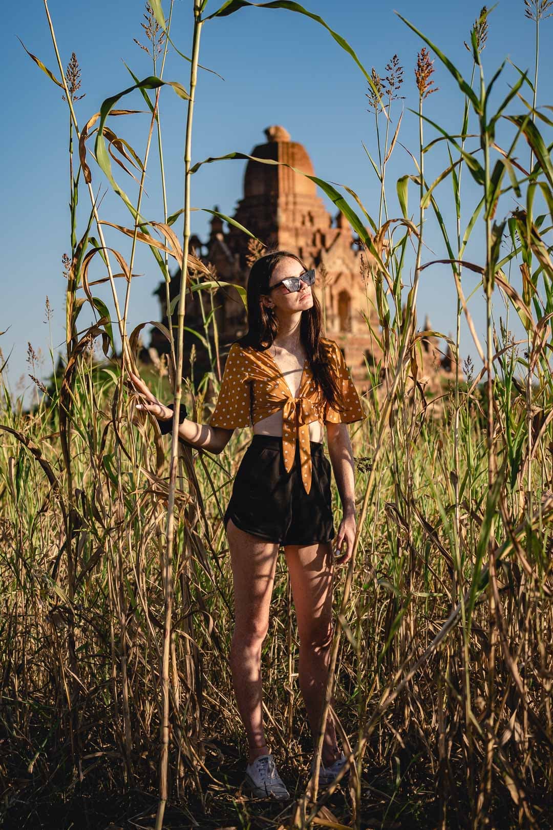 Jeune femme portant un haut moutarde noué et une mini-jupe noire, posant au milieu de hautes herbes devant un temple de Bagan sous un ciel bleu