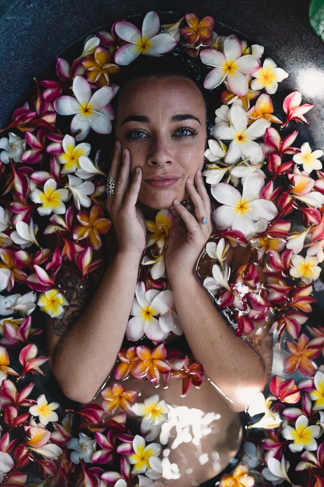 Portrait d'une femme immergée dans l'eau avec des fleurs de frangipanier roses, jaunes et blanches flottant autour de son visage