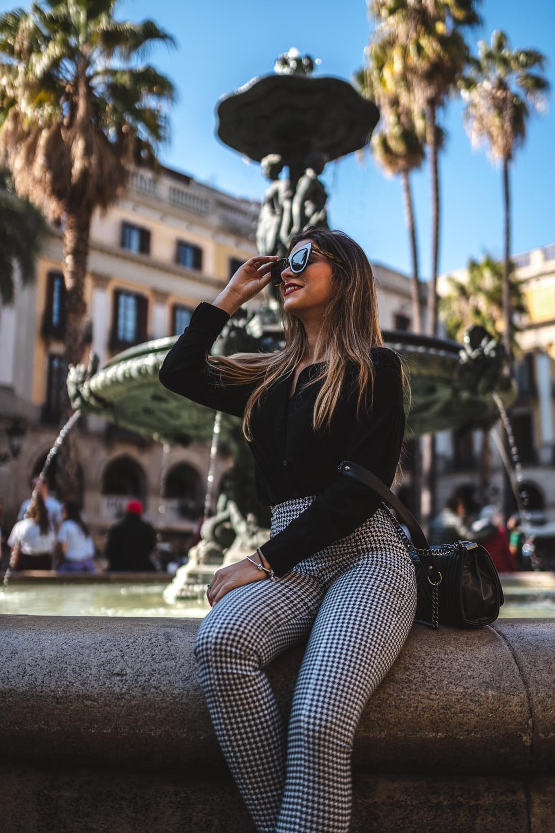 Femme portant des lunettes de soleil, un haut noir et un pantalon à motifs, assise sur le bord d'une fontaine à Barcelone