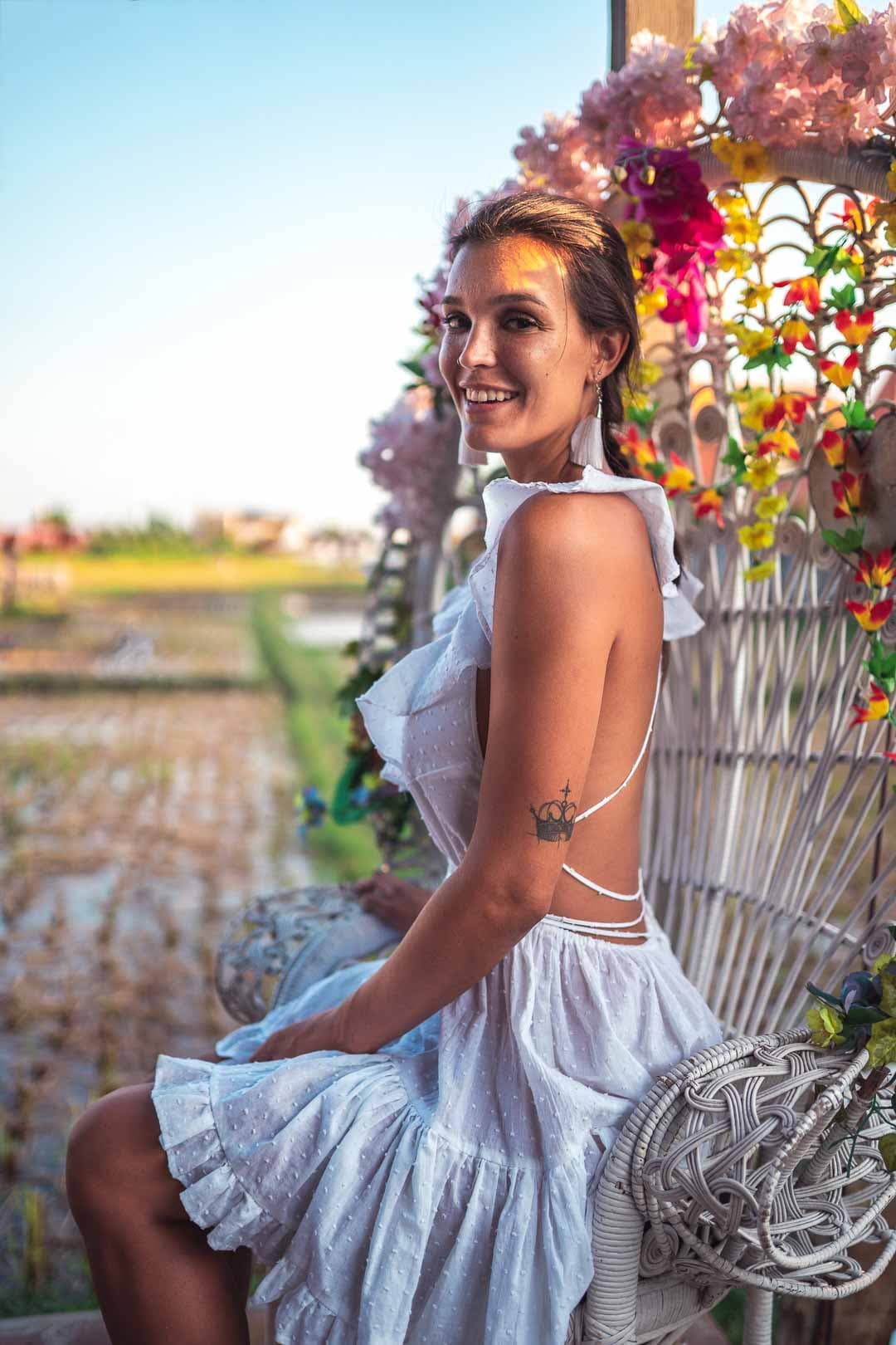 Femme portant une robe blanche à dos nu, assise sur un banc décoré de fleurs dans un champ de rizière avec un ciel bleu clair