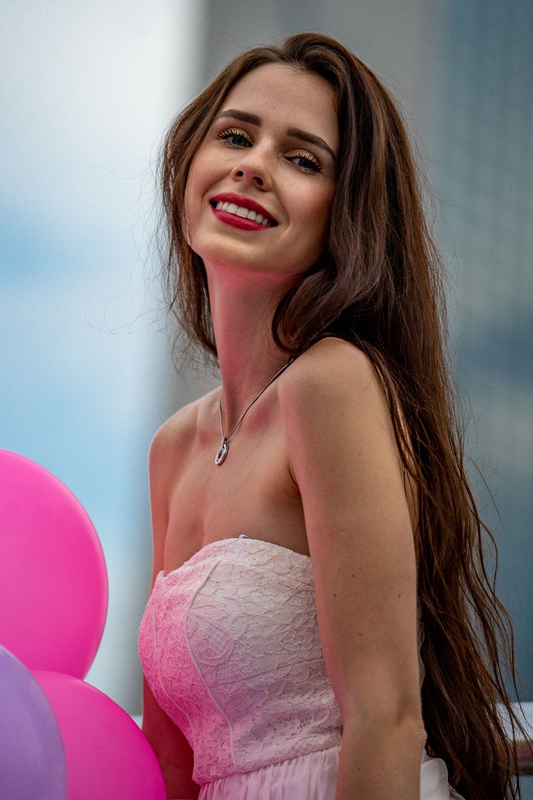 Portrait d'une femme souriante aux longs cheveux bruns, portant une robe bustier rose pâle et tenant des ballons roses