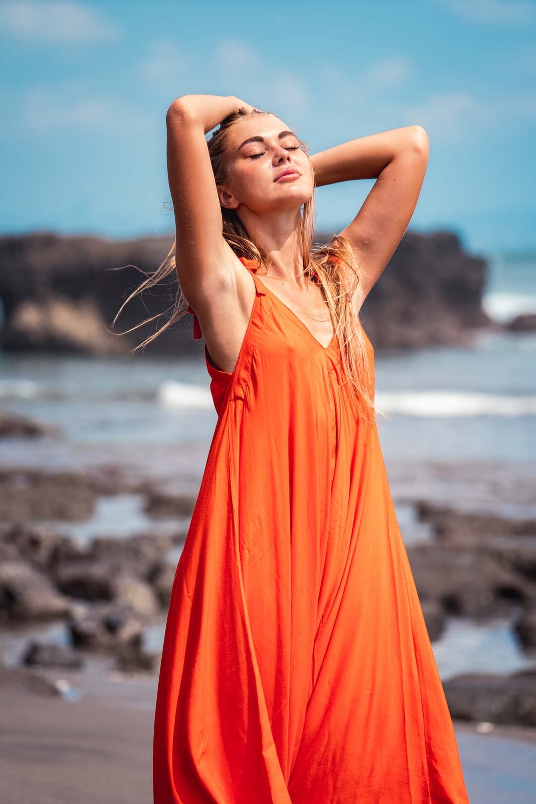 Jeune femme aux longs cheveux blonds, portant une robe longue orange, les bras levés sur une plage rocheuse avec la mer en arrière-plan