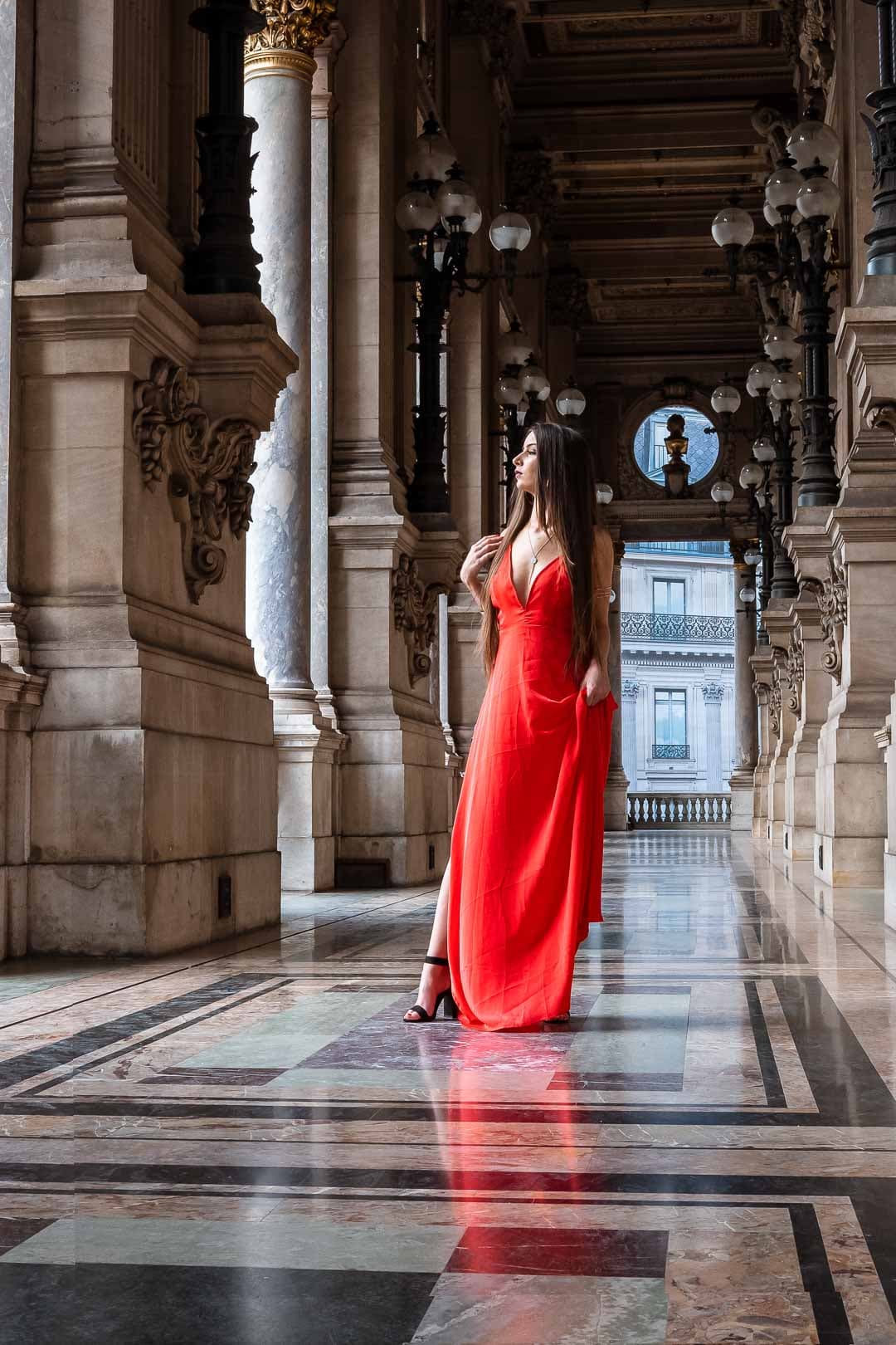 Femme élégante portant une longue robe rouge, marchant dans un long couloir avec des colonnes et une lumière douce