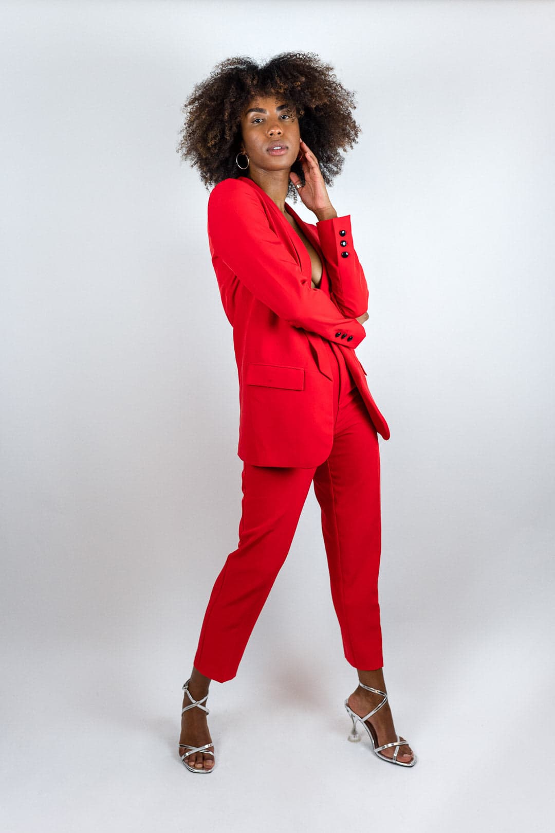 Femme en tailleur pantalon rouge vif, veste ouverte, main sur la nuque, cheveux afro, sandales argentées, studio fond gris clair