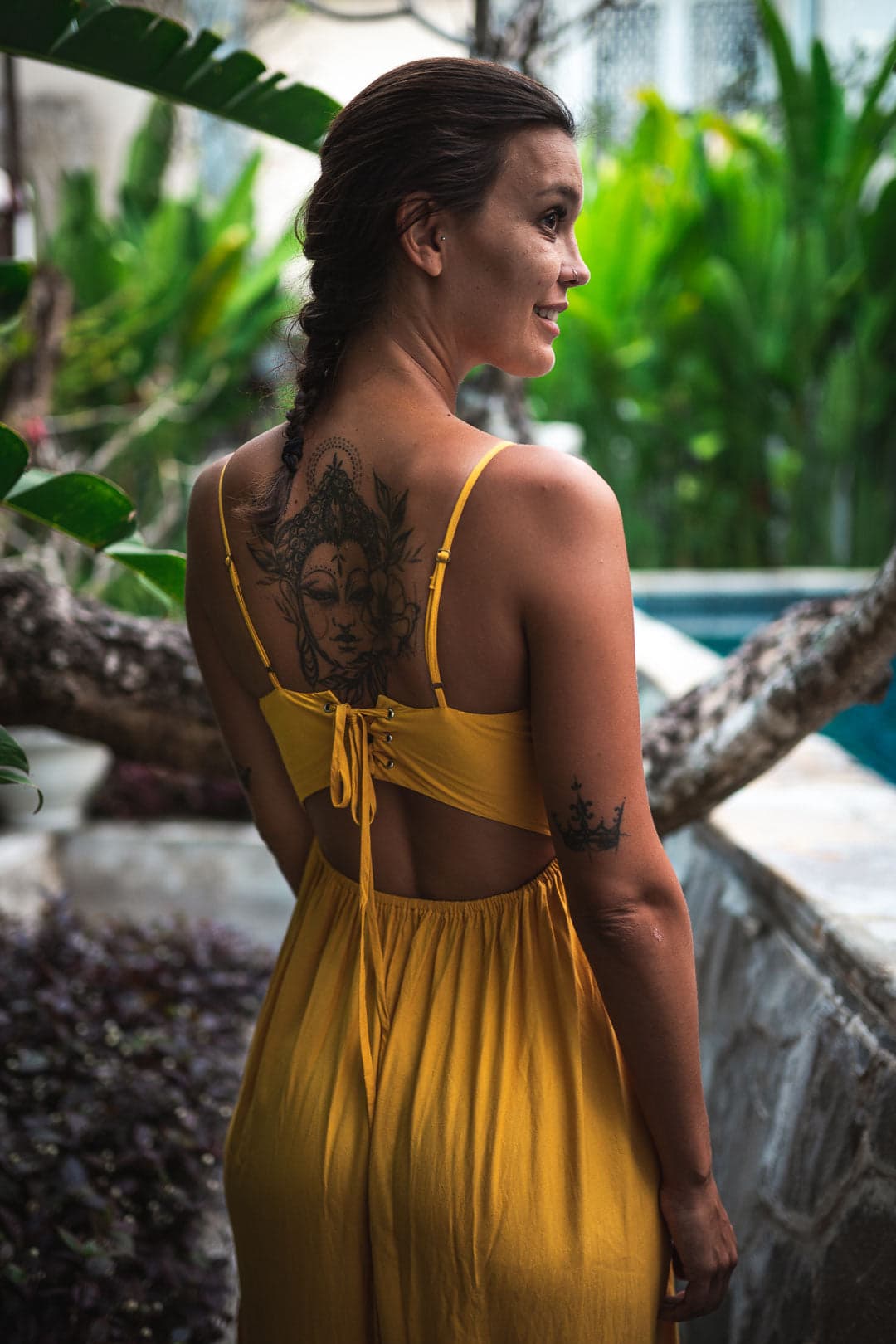 Jeune femme brune tatouée, de dos, portant une longue robe jaune à fines bretelles, regardant un paysage tropical avec une piscine