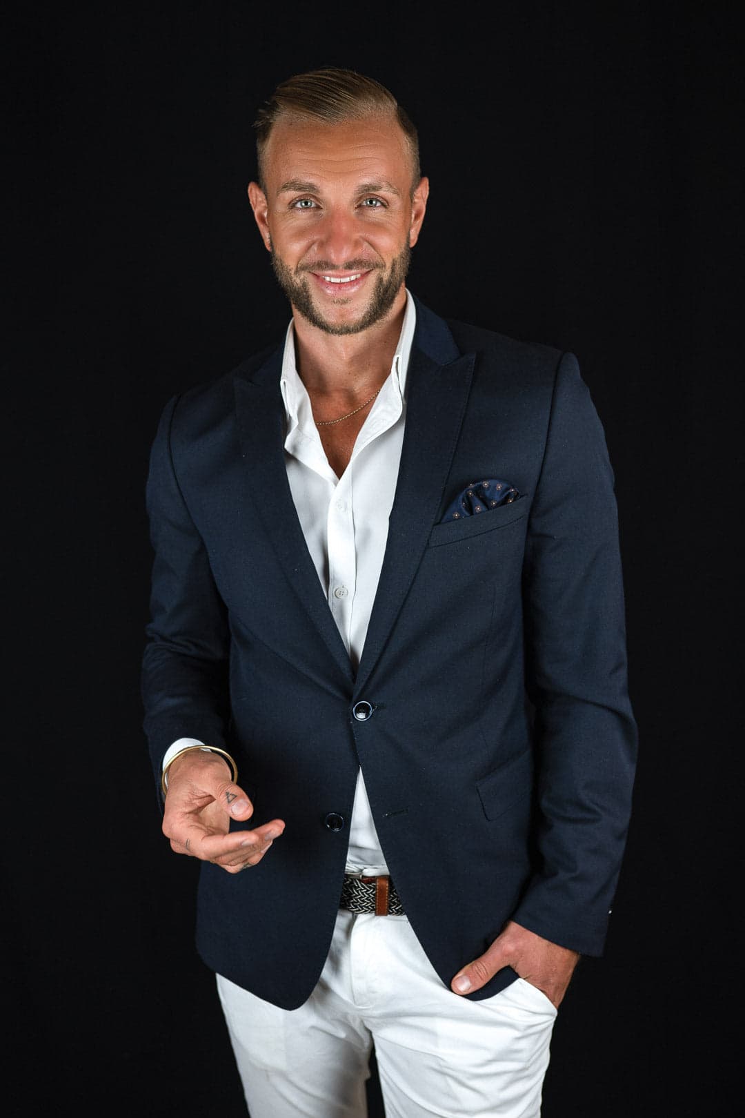 Portrait d'un homme souriant aux cheveux blonds coiffés en arrière, portant un blazer bleu marine sur une chemise blanche ouverte et un pantalon blanc, une main dans la poche