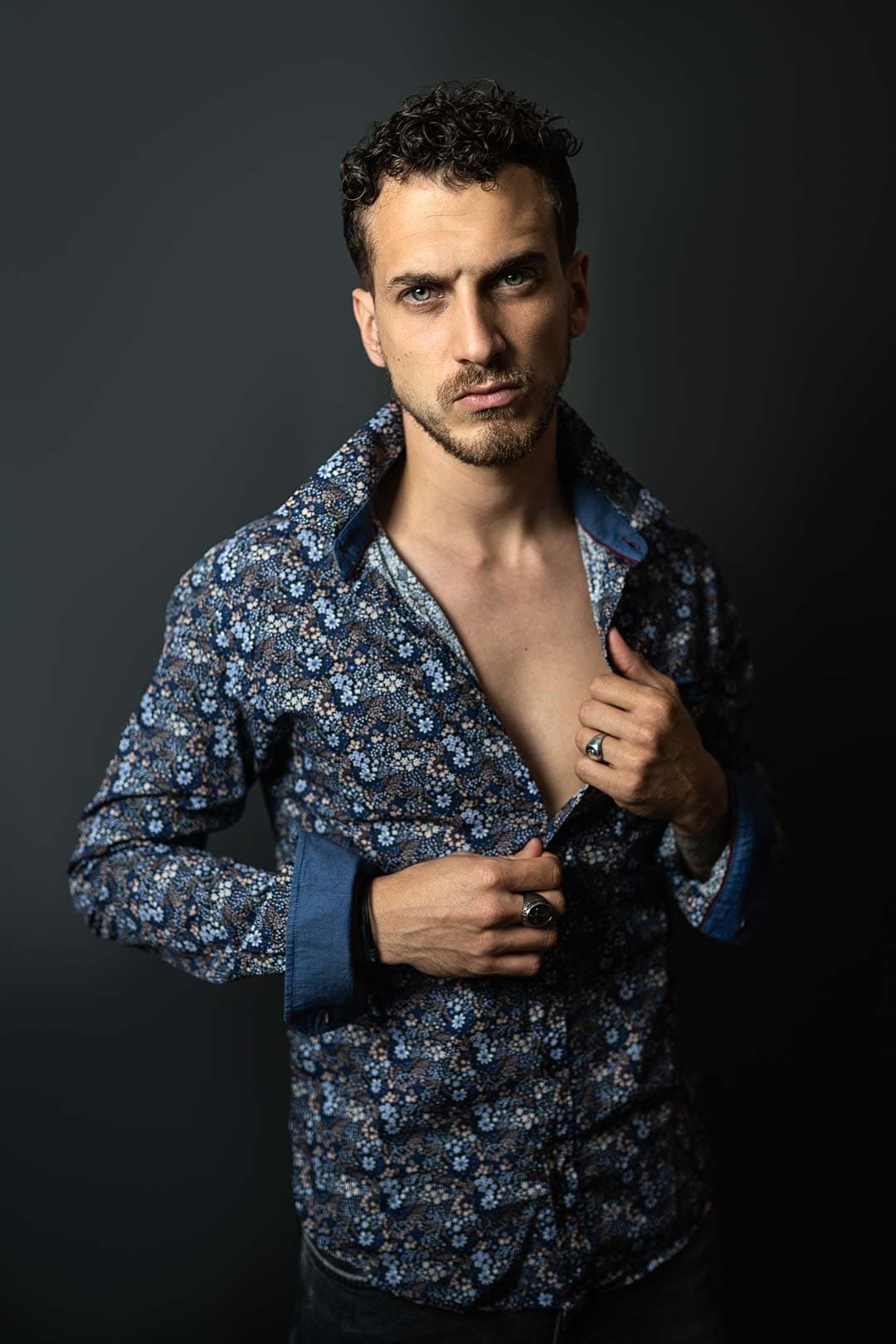 Portrait d'un jeune homme aux cheveux bruns bouclés, portant une chemise à motifs floraux bleus et blancs ouverte sur la poitrine