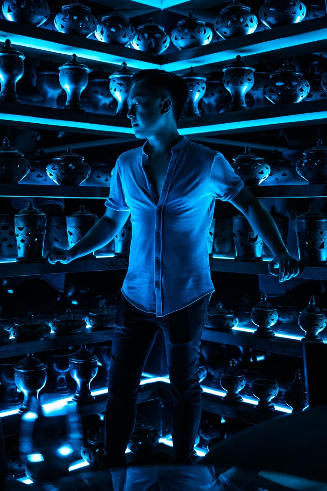 Homme portant une chemise blanche ouverte et un pantalon noir, debout dans une pièce futuriste éclairée par des lumières bleues