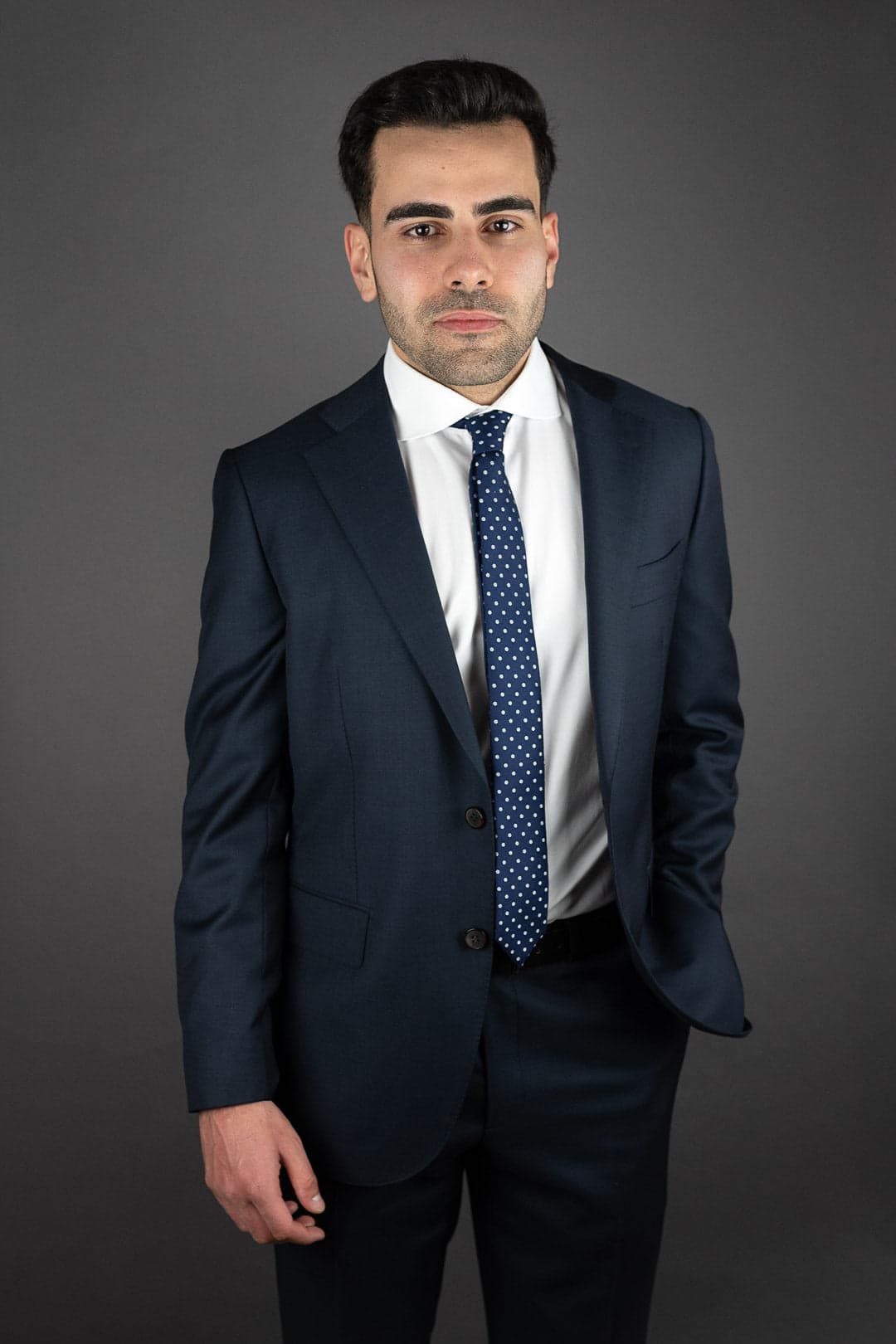 Portrait d'un jeune homme en costume bleu marine, chemise blanche et cravate à pois bleus et blancs, une main dans la poche