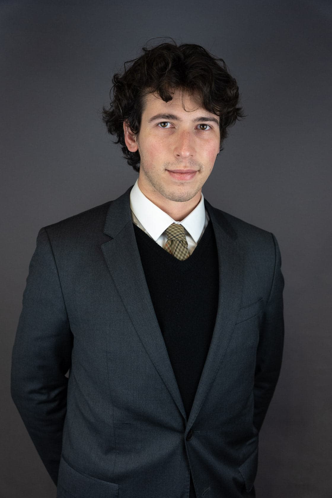 Portrait d'un jeune homme aux cheveux bruns bouclés, portant un costume gris foncé sur un pull noir et une chemise blanche avec une cravate à motifs