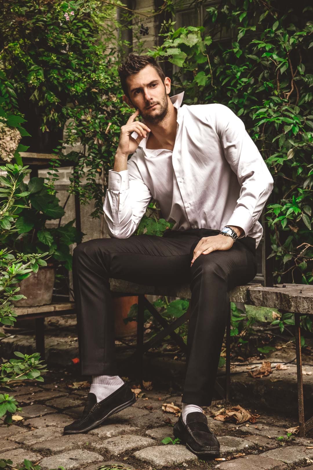 Homme élégant assis sur un banc en pierre dans un jardin luxuriant, portant une chemise blanche et un pantalon sombre