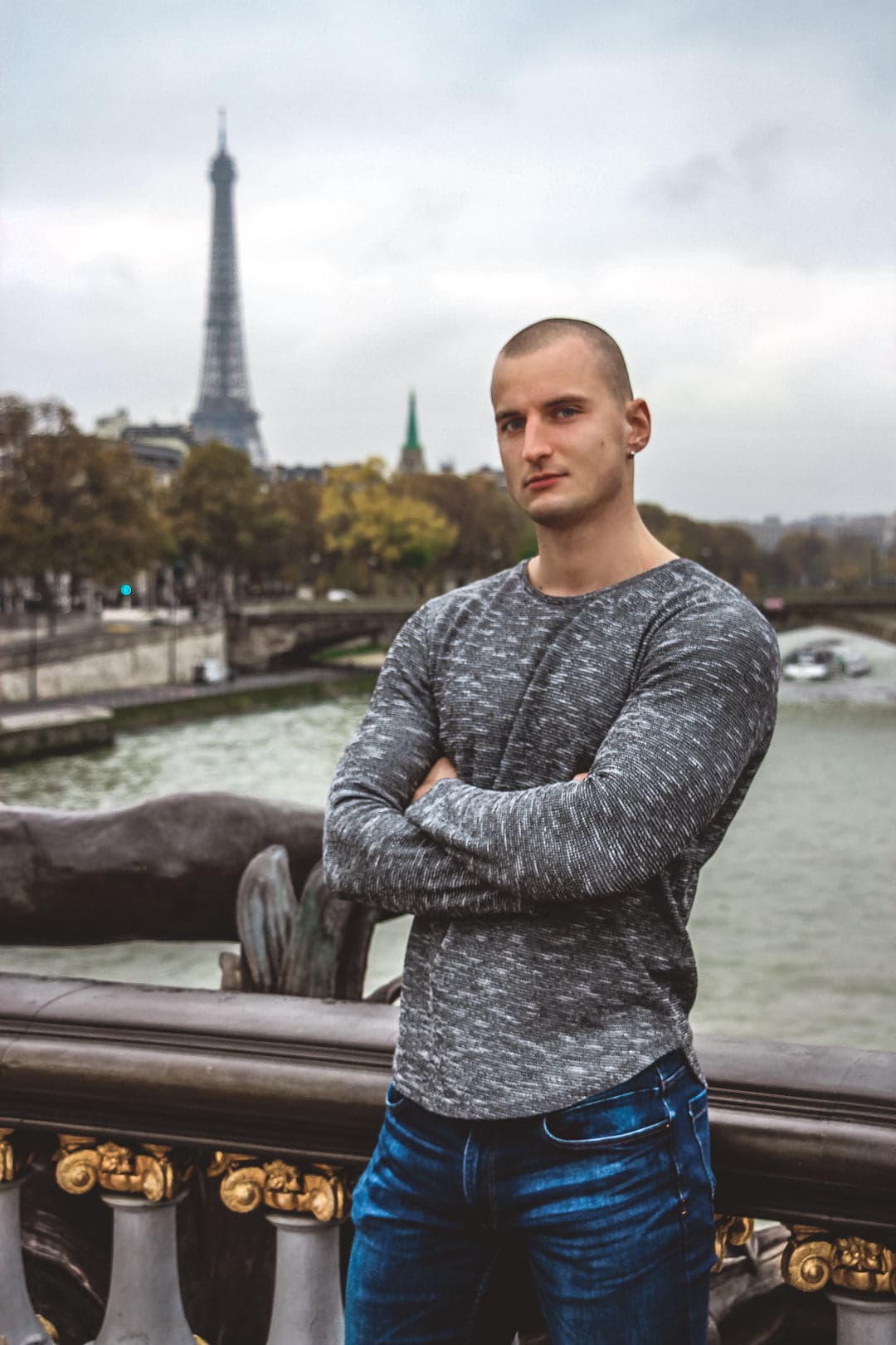 Homme chauve musclé portant un pull gris chiné et un jean, posant avec les bras croisés sur le pont Alexandre III avec la Seine et la Tour Eiffel en arrière-plan à Paris