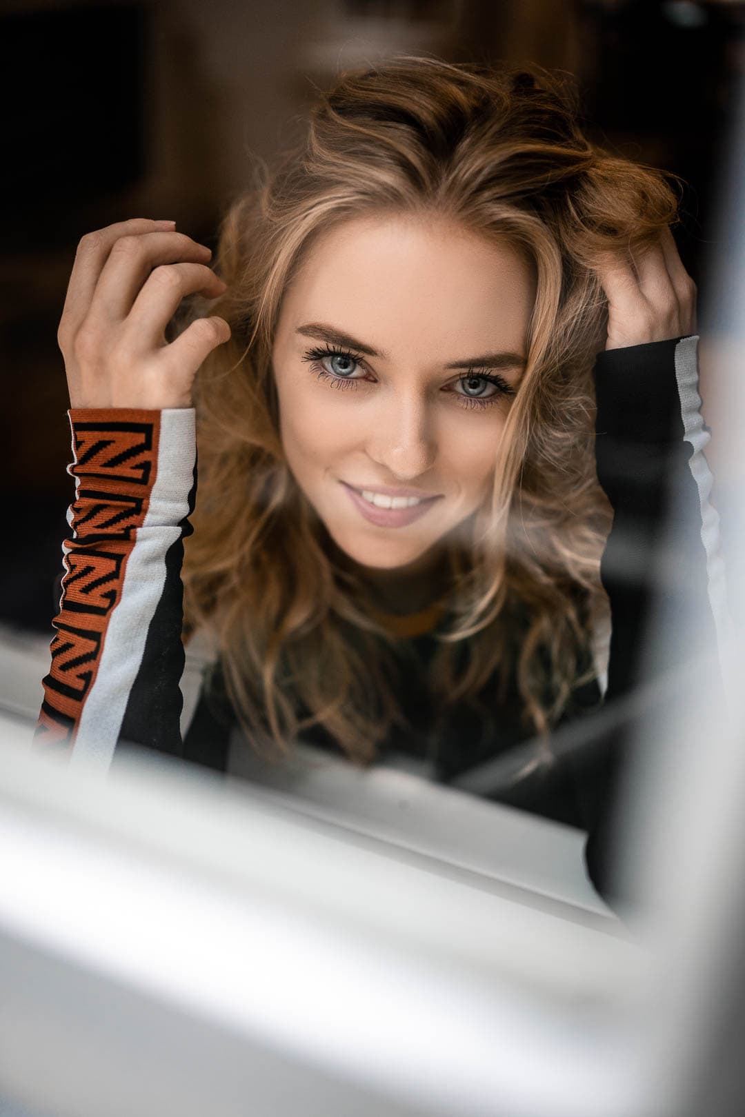 Portrait d'une jeune femme blonde souriante, les mains dans ses cheveux, regardant la caméra à travers une vitre