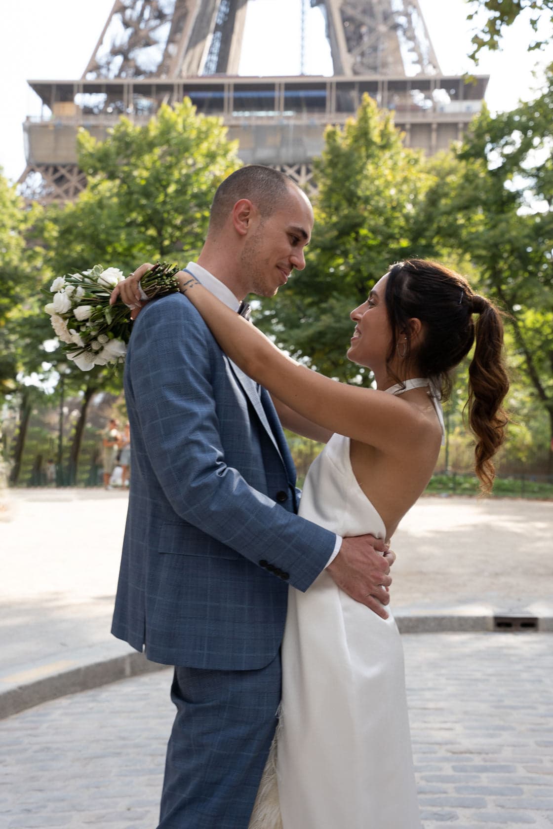 Un couple en tenue de mariage s'enlace devant la Tour Eiffel, la mariée tenant un bouquet de fleurs blanches