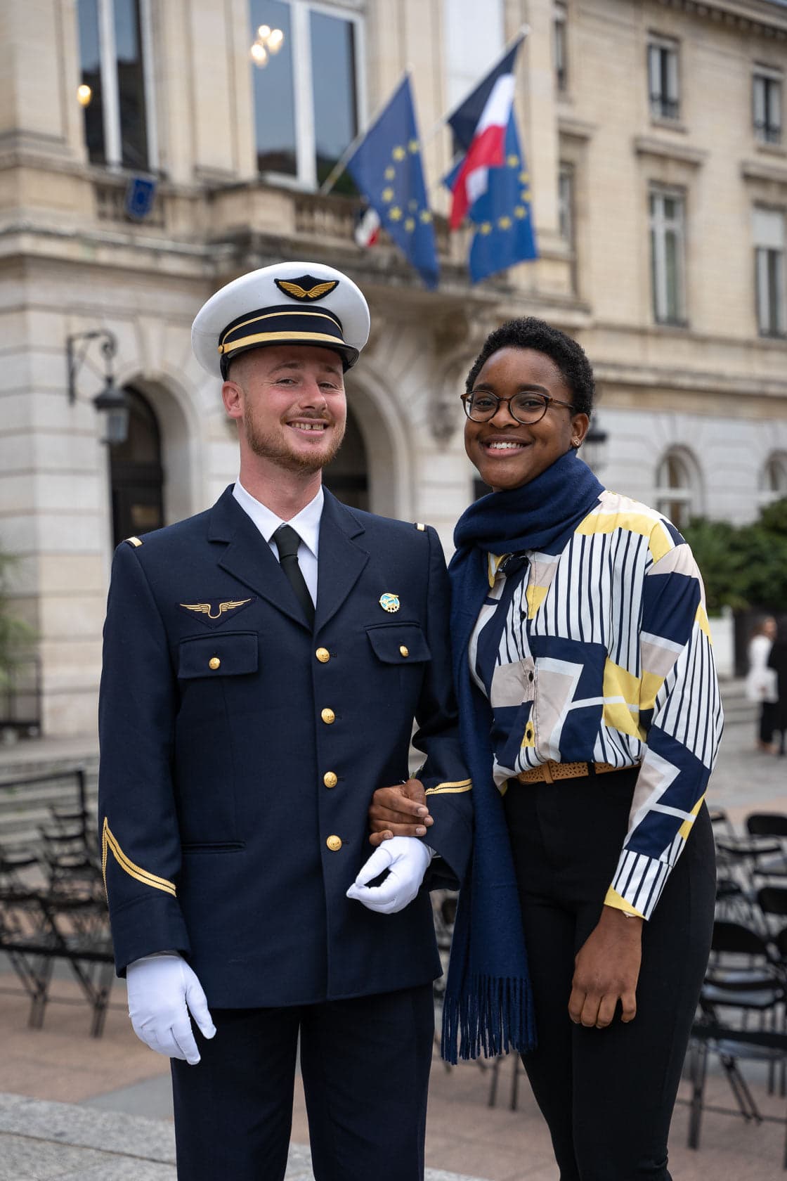 Un homme en uniforme militaire bleu marine avec des insignes dorés et une femme en haut géométrique blanc, bleu et jaune, portant un foulard bleu, se tiennent bras dessus bras dessous devant un bâtiment officiel avec des drapeaux français et européen