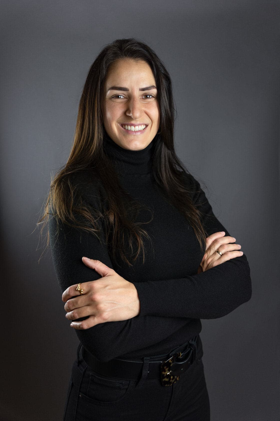Portrait d'une jeune femme souriante aux longs cheveux bruns, portant un pull à col roulé noir, bras croisés