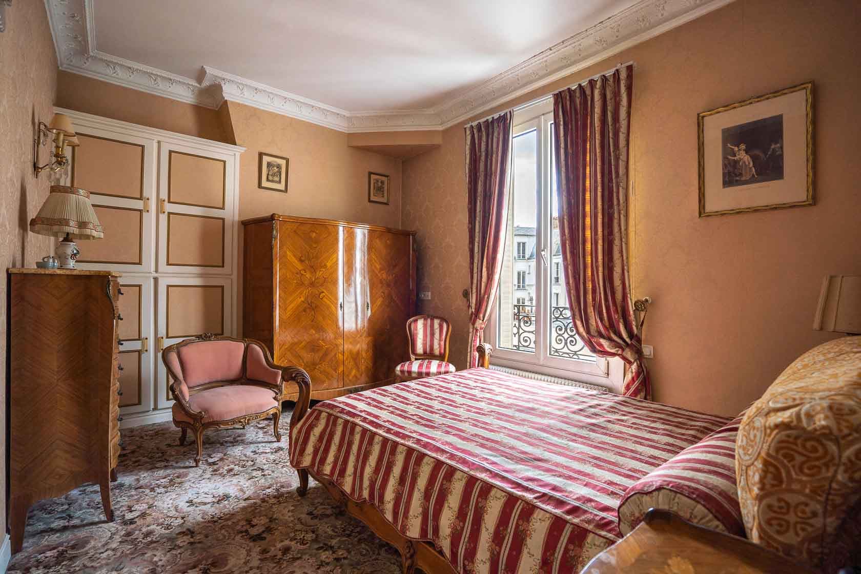 Chambre à coucher de style classique avec un lit rayé rouge et blanc, un grand meuble en bois, des fauteuils anciens et une fenêtre avec vue