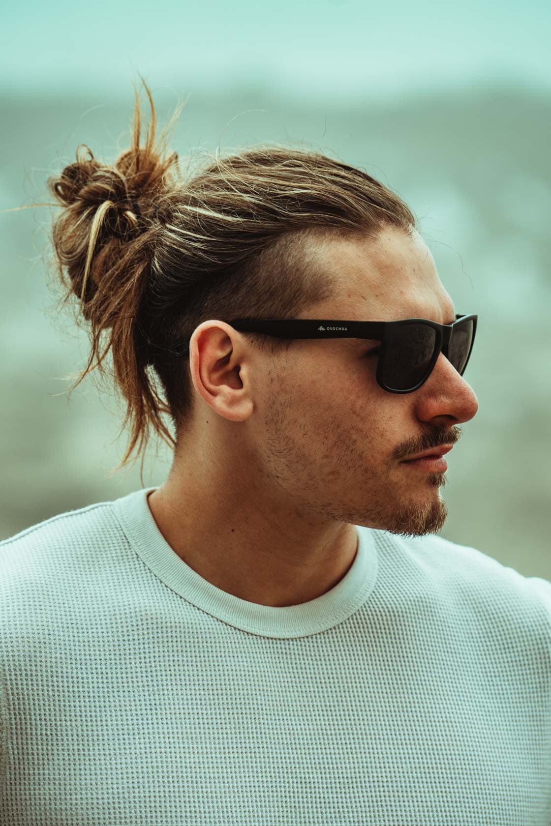 Portrait en extérieur d'un homme avec un chignon décontracté et des lunettes de soleil noires, regardant sur le côté avec une expression sereine