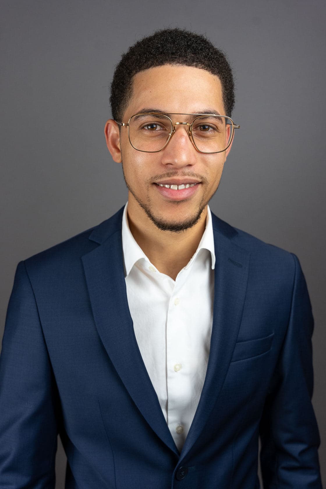 Portrait d'un jeune homme métissé souriant portant des lunettes à monture dorée, un blazer bleu marine et une chemise blanche