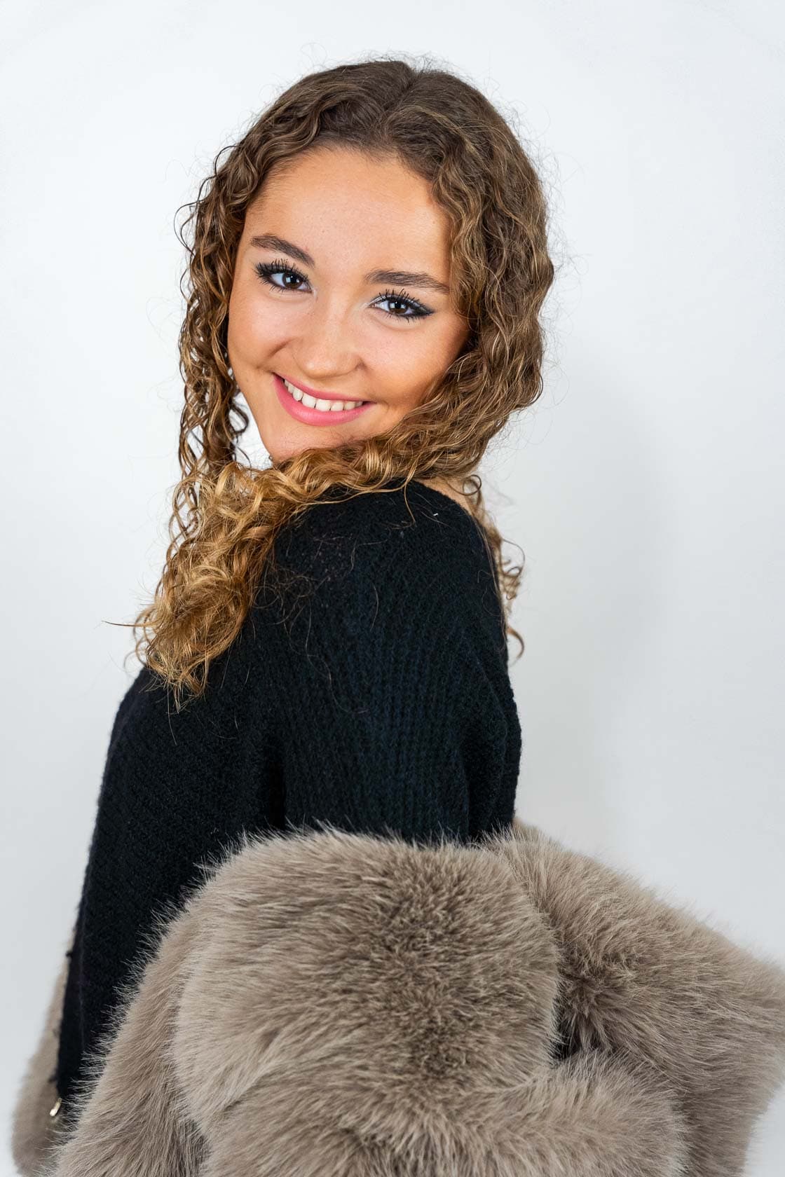 Portrait d'une jeune femme souriante aux cheveux bruns bouclés, portant un pull noir et une veste en fourrure grise sur l'épaule