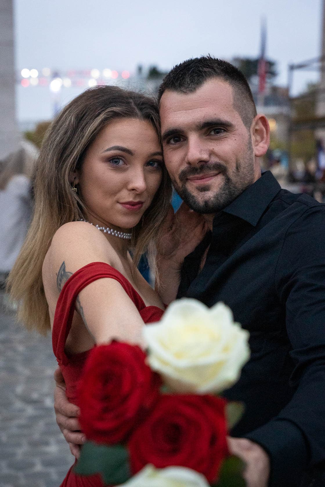 Un homme en chemise noire et une femme en robe rouge tenant un bouquet de roses rouges et blanches, enlacés sur le parvis de l'Arc de Triomphe lors d'une séance photo romantique
