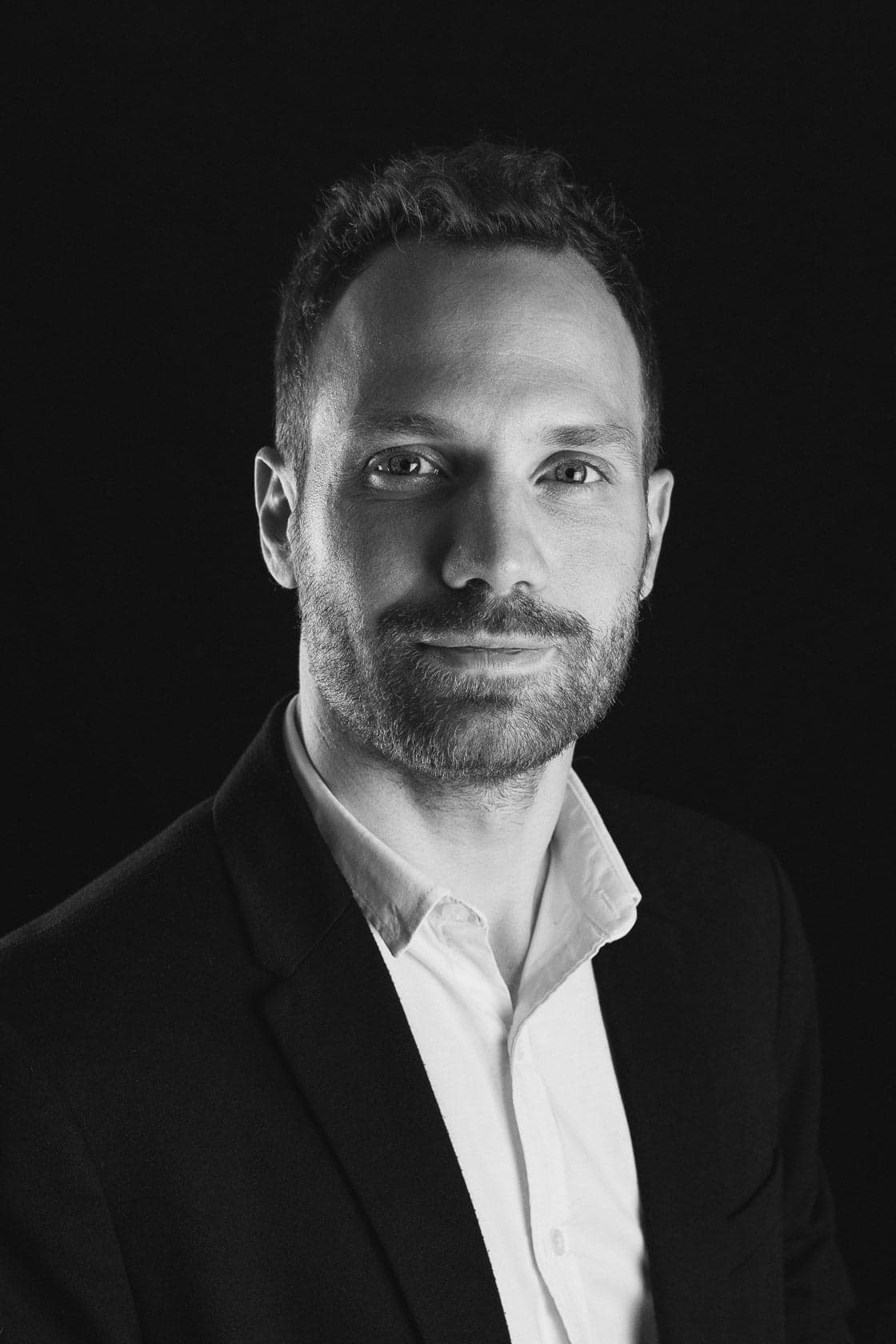 Portrait en noir et blanc d'un homme souriant avec une barbe soignée, portant un costume sombre sur une chemise claire