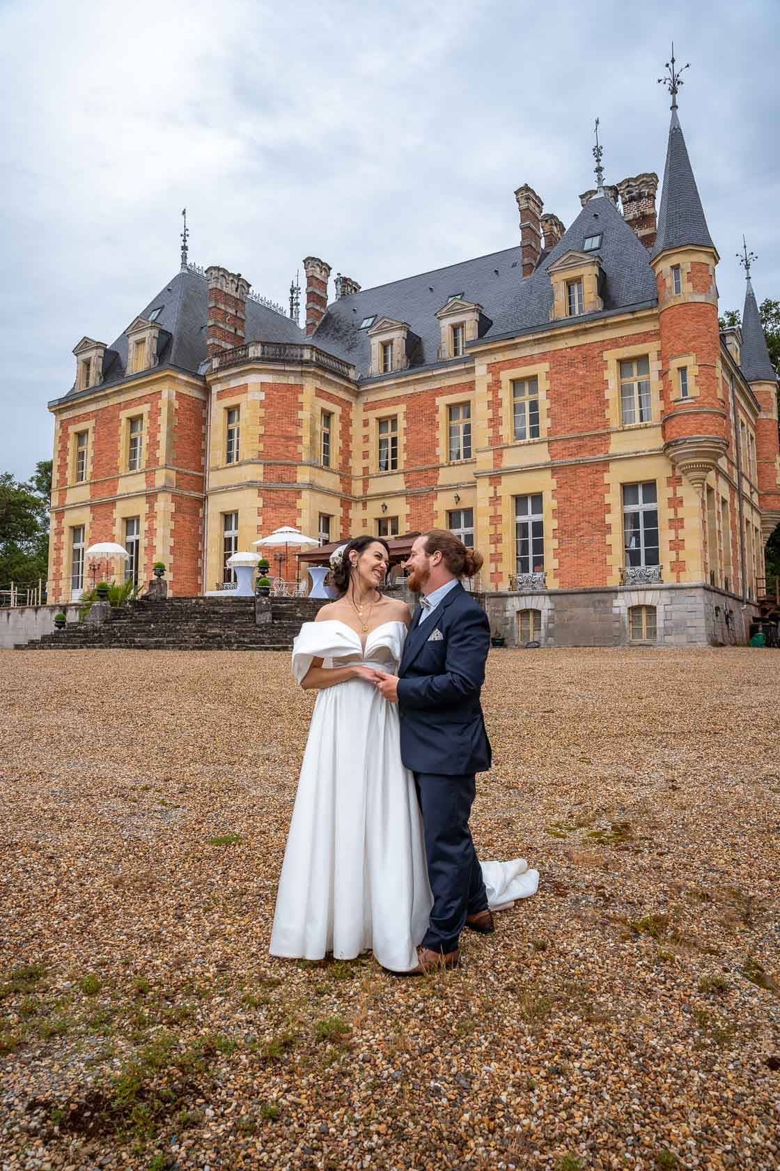 Les mariés posant devant un château en pierre et briques rouges, capturant un moment romantique sur le parvis